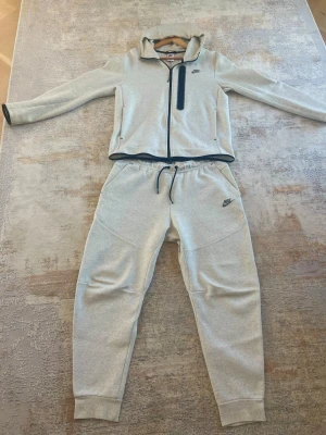Nike grå tech fleece - Säljer en Tech Fleece som tyvärr inte kommer till användning längre. Setet är endast använt ett fåtal gånger och är i mycket fint skick, i princip som nytt.  Tröjan är i storlek M och byxorna i storlek L.  Pris kan diskuteras vid snabb och smidig affär. Hör gärna av dig vid intresse eller om du har några frågor!