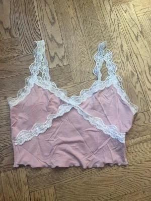 Rosa spetsbody med vita detaljer - Söt rosa body med vita spetsdetaljer längs axelband och urringning. Mjuk och stretchig modell med vågig kant nedtill. Perfekt för dig som gillar romantisk och feminin stil.