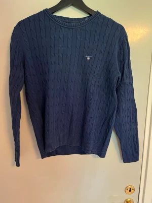 Marinblå kabelstickad tröja Gant - Kabelstickad marinblå tröja från Gant med rund halsringning och diskret logga broderad på bröstet. Tröjan har långa ärmar och ribbade muddar vid ärmslut och nederkant. Perfekt för en klassisk och stilren look.