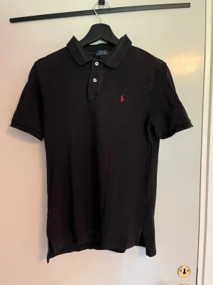 Ralph Lauren pike  - Klassisk svart pikétröja från Polo Ralph Lauren med korta ärmar och krage. Tröjan har knappar framtill och den ikoniska röda logotypen broderad på bröstet. Tillverkad i mjuk bomull för en bekväm passform.