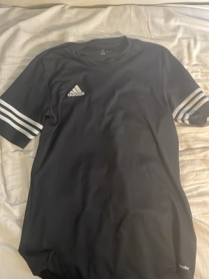 Svart Adidas t-shirt med vita ränder - Snygg svart t-shirt från Adidas med klassiska vita ränder på ärmarna och Adidas-logga på bröstet. Tillverkad i ett lätt och sportigt material som passar perfekt till en aktiv livsstil. Enkel och stilren design som funkar till det mesta.