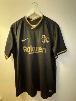 Svart FC Barcelona Messi tröja - Snygg svart FC Barcelona matchtröja från Nike med guldiga detaljer, klubbmärke och Rakuten-tryck på framsidan. På ryggen står det Messi och nummer 10 i guld. Tillverkad i lätt och ventilerande material, perfekt för fotboll eller träning.