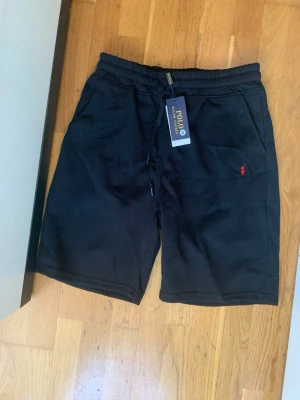 Svarta shorts från Polo Ralph Lauren - Svarta shorts från Polo Ralph Lauren med dragsko i midjan och klassisk röd logga broderad på ena benet. Tillverkade i mjuk bomull och har en avslappnad passform med fickor på sidorna. Perfekta för sommaren.