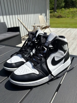 Air Jordan 1 mid svart/vit - Säljer ett par klassiska Air Jordan 1 Mid sneakers i svart och vitt läder. Skorna har den ikoniska swoosh-loggan på sidan, perforerad tåbox och högt skaft. Tidlös design med svarta skosnören och Air Jordan-logga på ankeln. Perfekta för dig som gillar streetwear och sneakers.