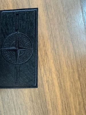 Stone island black badge - Säljer en svart rektangulär patch från Stone Island med det klassiska kompasslogotypen broderad ton-i-ton. Perfekt att fästa på jackor, väskor eller andra plagg för en stilren och exklusiv look.