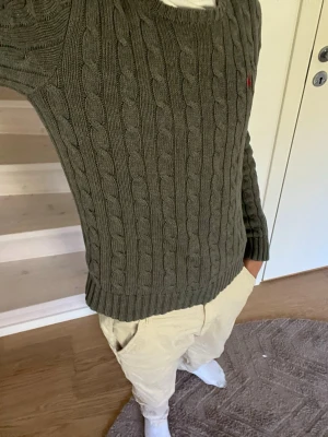 Olivgrön kabelstickad tröja - Säljer en olivgrön kabelstickad tröja med rund hals och ribbade muddar. Tröjan har en klassisk design med diskret logga på bröstet och är perfekt för lager-på-lager-stil. Materialet känns mjukt och värmande, perfekt för kyligare dagar.