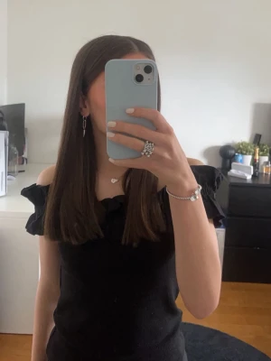 Svart offshoulder topp med volang - Snygg svart offshoulder topp med volangdetaljer längs axlarna. Toppen är ärmlös och har en croppad passform som ger en trendig look. Perfekt att styla med jeans eller kjol för en cool outfit.