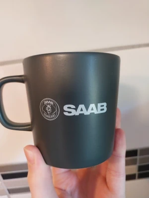 Svart/grå mugg med SAAB-logga - Snygg svart mugg i matt finish med SAAB-logga och texten 'SAAB' tryckt i vitt på sidan. Muggen har ett klassiskt handtag och är tillverkad i keramik, perfekt för kaffe eller te. Enkel och stilren design som passar både hemma och på jobbet. Oanvänd, odiskad, helt ny kopp med SAAB logga Saab Technologies Saab mugg Saab kaffekopp