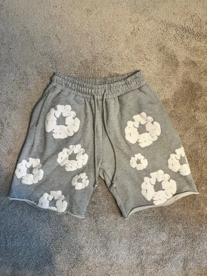 Denim Tears Shorts - Grå shorts med elastisk midja och snörning, dekorerade med stora vita blommönster över hela plagget. Mjukt material och avslappnad passform, perfekta för sommardagar. Shortsen har fickor både fram och bak. Strlk L
