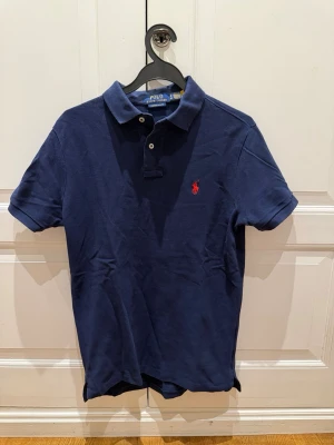 Mörkblå piké från Ralph Lauren - Klassisk mörkblå pikétröja från Ralph Lauren med röd broderad logga på bröstet. Tröjan har korta ärmar, krage och knappar framtill. Bomull