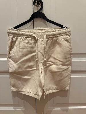 Beiga linne shorts med snörning - Snygga beiga linne shorts med elastisk midja och snörning framtill. Shortsen är ifrån A Days March och är i 100% linne. Storlek 46