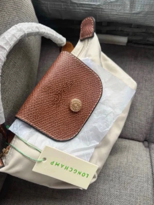 Beige handväska - Beige handväska från Longchamp med brunt läderlock och guldfärgad knapp. Väskan har ett klassiskt och tidlöst uttryck med praktiskt handtag och dragkedja. Tillverkad i slitstarkt tyg och detaljer i präglat läder. 🫶🏻