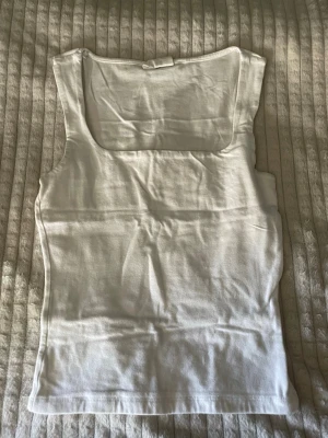 Vit linnetopp med fyrkantig ringning - Snygg vit linnetopp med breda axelband och fyrkantig ringning. Toppen är ärmlös och har en enkel, stilren design som passar perfekt till jeans eller kjol. Tillverkad i mjukt bomullsmaterial för en bekväm känsla hela dagen.