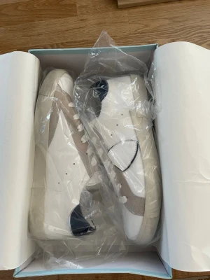 Vita sneakers från Philip Hog - Stilrena vita sneakers från Philip Hog med beige mockadetaljer och mörkblå häl. Skorna har vita snören och en klassisk låg siluett. Ovandelen är i läder och mocka, vilket ger en snygg kontrast. Perfekta för en clean och modern look.