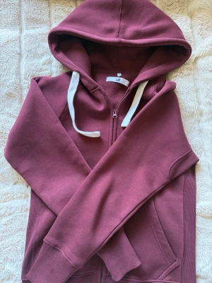 Vinröd hoodie med dragkedja - Vinröd hoodie med dragkedja och vita snören i huvan. Tillverkad i mjukt, fleeceliknande material på insidan. Klassisk passform med ribbade muddar och fickor framtill. Perfekt för en chill och avslappnad stil. Om man vill köpa separat så är det 400 för tröjan och 400 för byxorna 