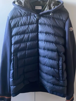 Mörkblå pufferjacka från Moncler - Snygg mörkblå pufferjacka från Moncler med vadderad framsida, mjuka ärmar och huva. Jackan har dragkedja, två fickor med dragkedja och Moncler-logga på ärmen. Perfekt för kyliga dagar och ger en sportig look, hör gärna av dig vid frågor:)