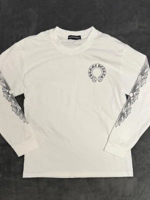 Chrome Hearts tröja - Knappt använd Vit långärmad topp från Chrome Hearts med svart tryck på bröstet och ryggen. Ärmarna har snygga grafiska detaljer längs sidorna. Tillverkad i mjuk bomull och har en normal passform. Perfekt för dig som gillar streetwear och unika prints. Pris kan diskuteras
