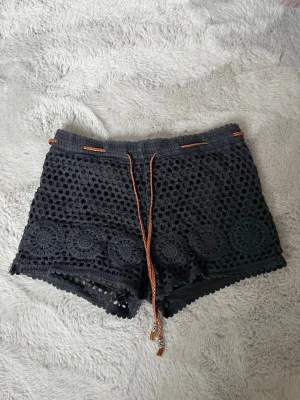 Svarta virkade shorts med snörning - Svarta virkade shorts med snyggt mönster och dekorativa cirklar längs nederkanten. De har en orange snörning i midjan och är fodrade för extra komfort. Perfekta för sommarens varma dagar och festivaler.