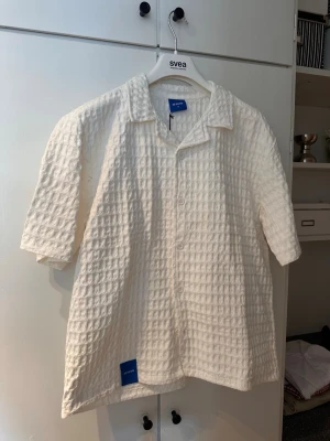 Vit skjorta - Vit skjorta med kort ärm och avslappnad passform. Skjortan har ett strukturerat, rutigt tyg i bomull och knäppning framtill. Perfekt för en chill och trendig look.