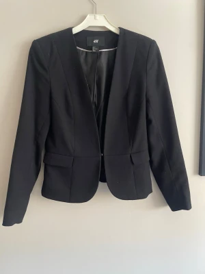 Svart kavaj från H&M - Stilren svart kavaj från H&M med figurnära passform och öppen front. Kavajen har långa ärmar, markerade axlar och dekorativa fickor framtill. Tillverkad i ett slätt tyg som ger en klassisk look.