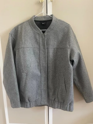 Grå ullrock med resår från H&M - Stilren grå rock i ullblandning från H&M med rak passform, ståkrage och dold dragkedja. Två snedställda fickor framtill och resår i nederkant ger en modern look. Perfekt för kyliga dagar.