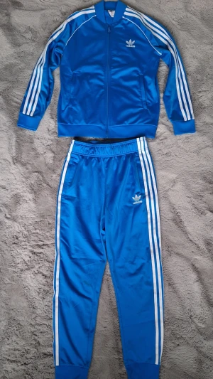 Adidasdress Adicolor SST Track set stl 158 - Stället är i mycket gott skick. Liten fläck från cykelkedja på ena benet, kolla bild 4.   Färg: Blue bird.  Normal passformSnygga blå träningsbyxor och träningsjacka från Adidas med klassiska vita ränder längs sidorna och Adidas-logga på låret. Byxorna har elastisk midja och är gjorda i glansigt syntetmaterial som ger en sportig vibe. Perfekta för chill eller träning.