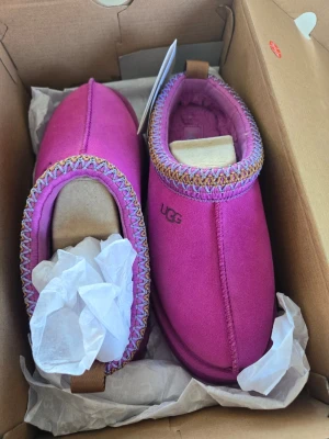 Rosa tofflor från UGG Taz  - Säljer ett par snygga rosa tofflor från UGG med färgglad brodyr runt öppningen. Tofflorna har en mjuk insida och en stadig sula, perfekta att glida runt i hemma. Materialet är mocka och de har en klassisk UGG-logga på sidan.