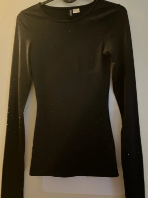 Svart glittrig långärmad topp H&M - Snygg svart långärmad topp från H&M med glittriga detaljer över hela tyget. Tight passform och rund halsringning, perfekt att styla med jeans eller kjol för en trendig look.