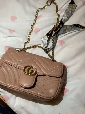 Beige axelväska från G - Säljer en snygg beige axelväska från G med guldfärgad kedja och GG-logo framtill. Väskan har quiltat mönster med hjärtdetalj på baksidan och en dekorativ scarf knuten på axelremmen. Perfekt för att lyfta din outfit. Men knappen som håller till där man stänger är sönder, så man kan egentligen inte stänga” väskan, därför följer den väldigt billigt annars funkar väskan som den ska:) och är väldigt fin