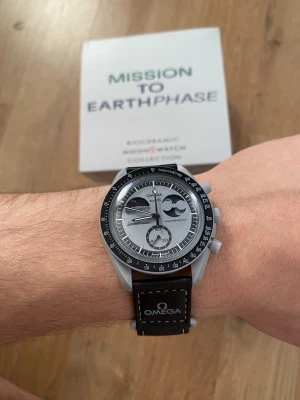 Omega x Swatch Mission to Earthphase - Snygg Omega x Swatch Mission to Earthphase från Bioceramic Moonswatch Collection. Klockan har en silverfärgad urtavla med svarta detaljer, tachymeter och svart läderband med Omega-logga. Perfekt för dig som gillar stilrena accessoarer med unik design.