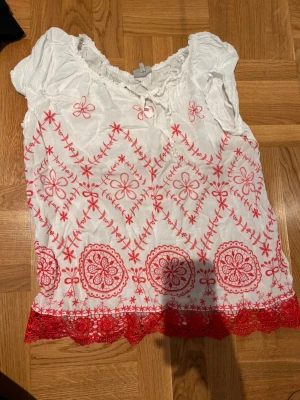 Vit blus med röd brodyr och spets - Söt vit blus med röda broderade blommor och mönster, med röd spetskant nertill. 