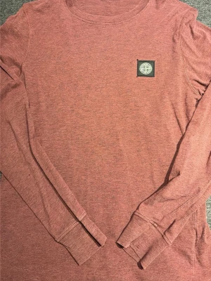 Röd långärmad tröja Stone Island - Säljer en snygg långärmad tröja från Stone Island i en dämpad rosa nyans. Tröjan har rund halsringning och en ikonisk logotyp-patch på bröstet. Tillverkad i mjukt bomullsmaterial som känns skönt mot huden.