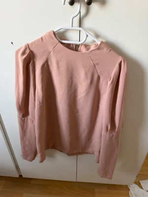 Ljusrosa blus med lång ärm - Säljer en stilren ljusrosa blus med rund halsringning och långa ärmar. Blusen har en diskret dragkedja bak och lätt puff vid axlarna för en trendig look. Materialet känns mjukt och har ett fint fall. Perfekt att matcha med jeans eller kjol.