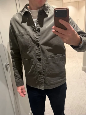 Grön overshirt från J.Lindeberg - Stilren grön overshirt från J.Lindeberg i kraftig bomull. Klassisk krage, tre stora fickor framtill och knäppning med svarta knappar. Perfekt lager-på-lager-plagg med avslappnad passform och tidlös design.