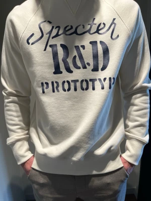 Vit sweatshirt från Specter Design - Snygg vit sweatshirt med blå texttryck 'Specter R&D PROTOTYP' på bröstet. Tröjan har rund hals, långa ärmar och ribbade muddar. Perfekt för en avslappnad och trendig stil. Helt oanvänd 