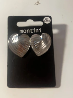 Silverfärgade hjärtörhängen Montini - Snygga örhängen i form av hjärtan från Montini. De är silverfärgade med räfflad yta som ger en cool och trendig look. Perfekta för att lyfta din outfit med en söt detalj. Stiftmodell med plastbaksida.