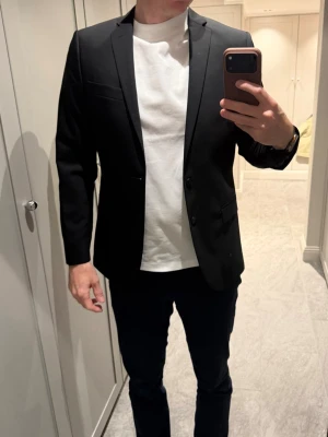 Svart kostym stl48 J.Lindeberg  - Säljer en stilren svart kostym med slimmad passform och snygga detaljer som knappar på ärmsluten och två framfickor. Kavajen har en bröstficka och är tillverkad i ett slätt material som ger ett elegant intryck. Perfekt för dig som vill ha en tidlös look. Modell: Mick.