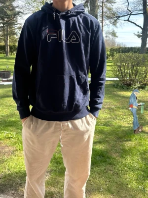 Mörkblå hoodie från FILA - Säljer en mörkblå hoodie från FILA med stor logga i vitt, rött och blått på bröstet. Hoodien har huva med dragsko och en stor magficka. Materialet är mjukt och bekvämt, perfekt för chill dagar. Långa ärmar och ribbade muddar vid ärmslut och nederkant.