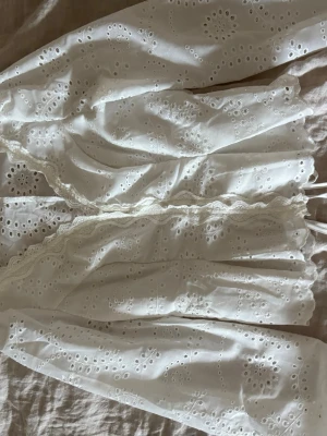 Vit broderad blus med spetsdetaljer - Söt vit blus med broderade hålmönster och romantiska spetsdetaljer längs knäppningen. Blusen har långärmade ärmar och är gjord i ett lätt bomullstyg som känns luftigt och fräscht. Perfekt för dig som gillar en feminin och bohemisk stil.