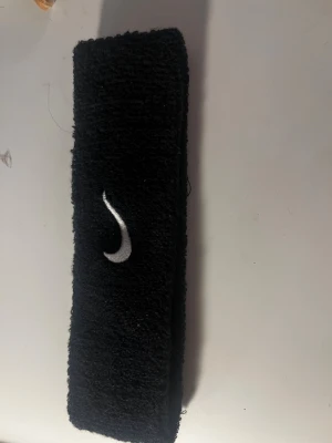 Svettband från Nike i svart - Svart svettband från Nike med broderad vit swoosh-logga. Tillverkat i mjukt och stretchigt frottématerial som absorberar svett effektivt. Perfekt för träning, gym eller sportaktiviteter.