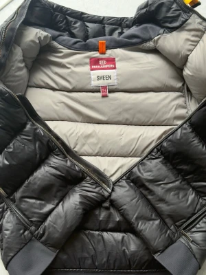 Svart pufferjacka Parajumpers Sheen - Snygg svart pufferjacka från Parajumpers, modell Sheen. Jackan har quiltad design, dragkedja framtill och breda muddar vid ärmslut och nederkant. Insidan är ljusgrå och jackan har en hög krage. Perfekt för kalla vinterdagar.