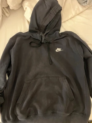 Svart Nike hoodie med logga - Svart hoodie från Nike med vit broderad logga på bröstet. Klassisk känguruficka framtill och justerbar huva med snören. Mjuk bomullsblandning som är skön att ha på sig. Perfekt för en avslappnad och sportig stil.
