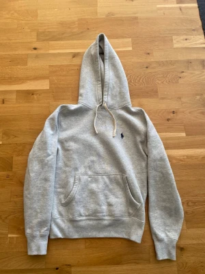 Grå hoodie från Polo Ralph Lauren - Klassisk grå hoodie från Polo Ralph Lauren med broderad marinblå logga på bröstet. Hoodien har huva med dragsko, magficka och ribbade muddar vid ärmslut och nederkant. Perfekt för en avslappnad och stilren look. Storlek Xs passar under 170, 