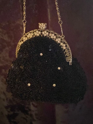 Svart pärlbroderad kuvertväska - Unik svart kuvertväska med pärlbroderi och glittrande stenar. Väskan har en elegant metallram med dekorativa detaljer och en tunn kedja som axelrem. Perfekt accessoar för att lyfta din outfit med lite extra bling och vintagekänsla.