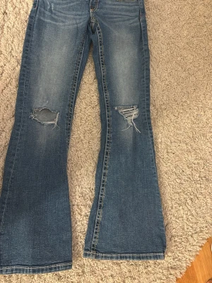 Blå bootcut jeans med slitningar - Säljer ett par blå bootcut jeans från Gina tricot med slitna detaljer på båda benen. Jeansen har klassisk femficksdesign, synliga sömmar i kontrastfärg och dragkedja med knapp. 