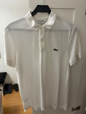 Vit piké Lacoste - Klassisk vit pikétröja från Lacoste med korta ärmar och krage. Tröjan har slim fit-passform, knäppning framtill och den ikoniska gröna krokodilloggan broderad på bröstet. Perfekt för en clean och sportig stil.