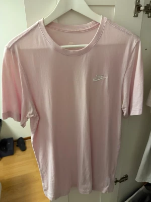 Ljusrosa Nike t-shirt i bomull - Snygg ljusrosa t-shirt från Nike med klassisk logga på bröstet. T-shirten har rund halsringning och är tillverkad i mjuk bomull för en skön känsla. Perfekt för en avslappnad och sportig stil.