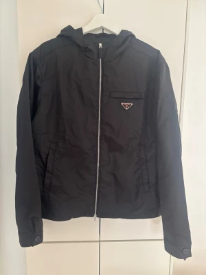 Svart vindjacka från Prada - Snygg svart vindjacka från Prada med dragkedja framtill och huva. Jackan har en bröstficka med det klassiska Prada-emblemet i metall och två sidofickor. Tillverkad i ett lätt och slitstarkt material som passar perfekt för blåsiga dagar. Storlek på jackan är 46 som motsvarar S-M. Jackan är helt ny! 