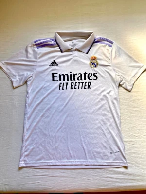Real Madrid matchtröja Adidas M - Snygg vit Real Madrid matchtröja från Adidas med lila detaljer på axlarna och klubbmärke på bröstet. Klassisk krage och svart Adidas-logga samt Emirates Fly Better-tryck på framsidan. Tillverkad i lätt och funktionellt material.
