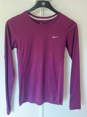 Lila långärmad träningströja Nike - Snygg lila långärmad träningströja från Nike med Dri-FIT-material som andas. Tröjan har rund halsringning och en liten vit Nike-logga på bröstet. Perfekt för löpning eller gympass.
